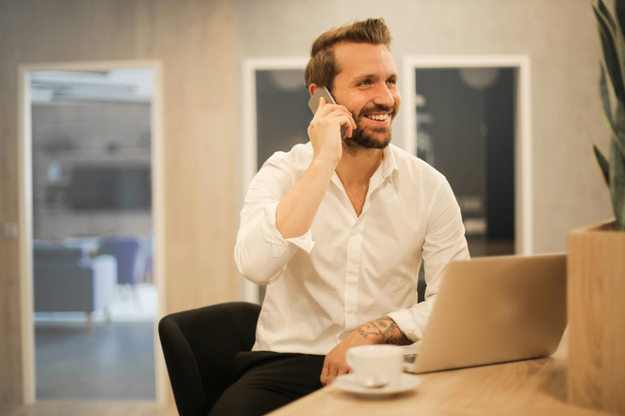 A smiling man in a white shirt talks on his phone while sitting at a desk with a laptop and coffee cup in a modern office setting.