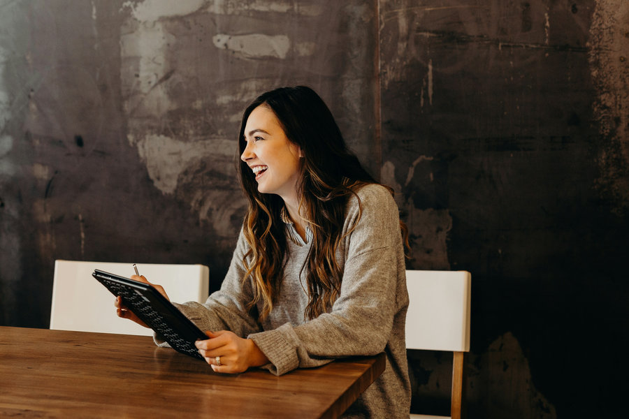 A woman with long brown hair wearing a grey sweater sits at a wooden table, smiling and holding a tablet with a stylus, in front of a dark textured wall